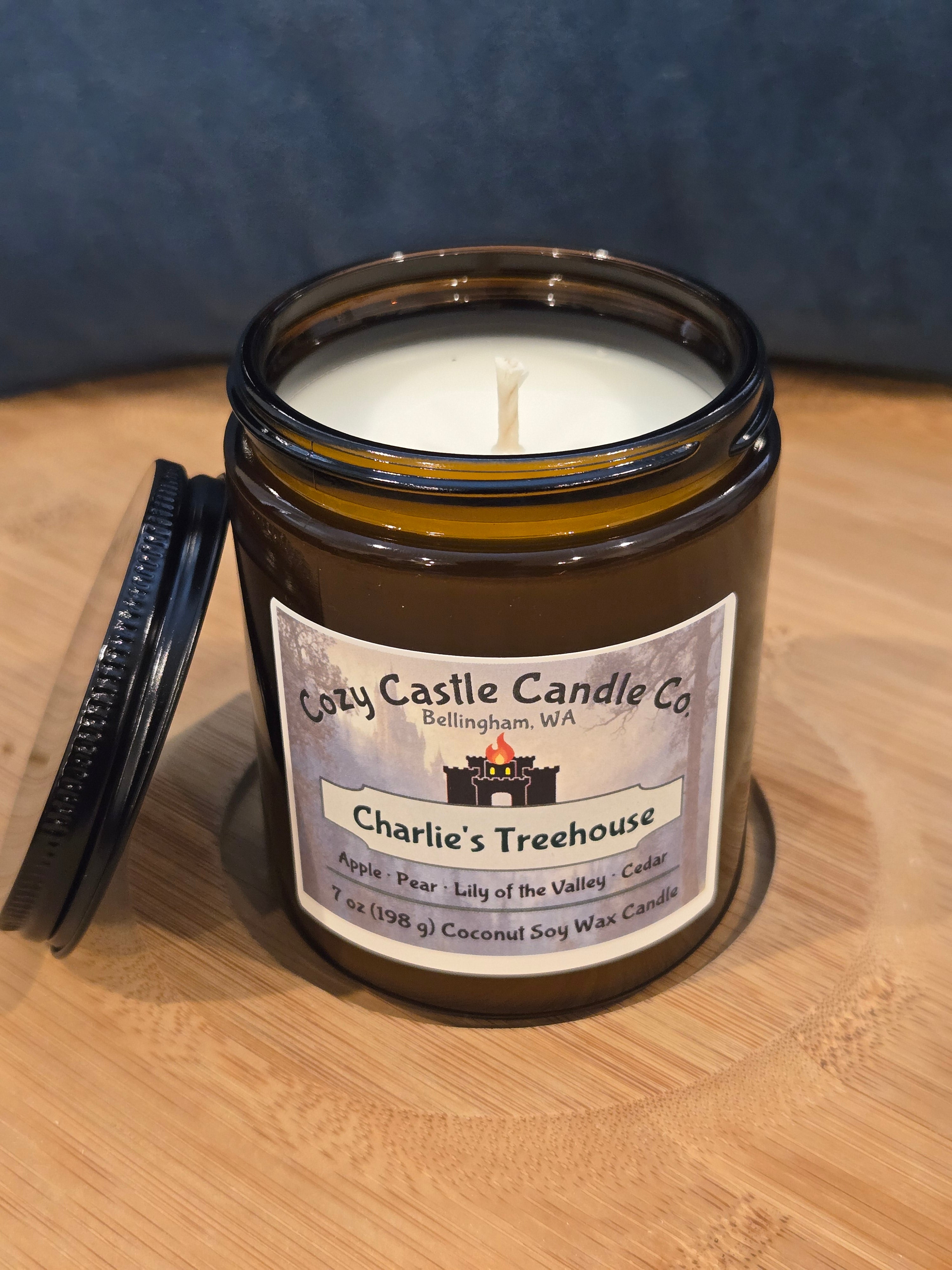Candle in a jar with a label on a wooden surface. Scent: Charlies Treehouse, with Apple, Pear, Lily of the Valley, and Cedar.