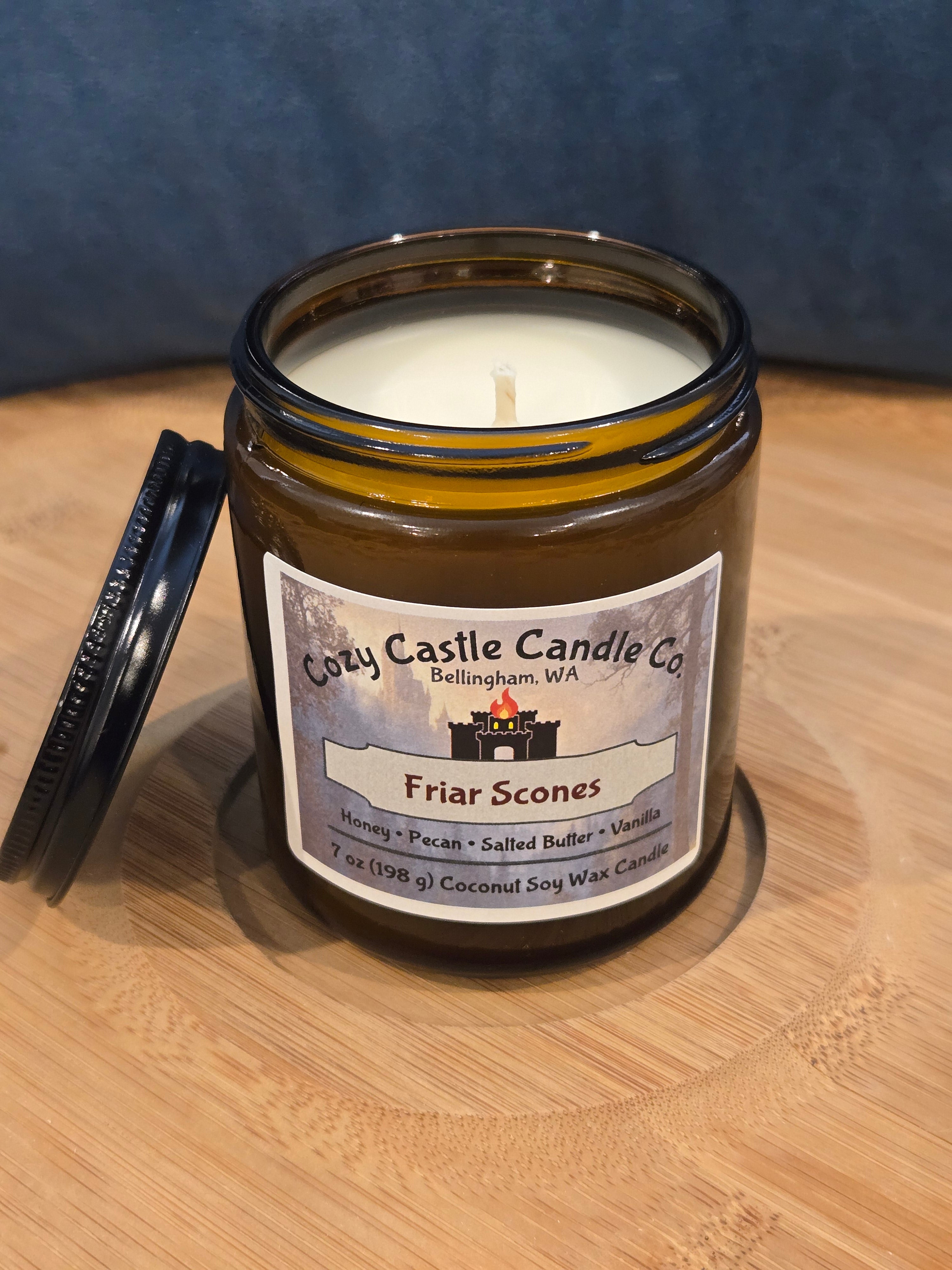Candle in a glass jar with a label on a wooden surface. Friar Scones, with notes of Honey, Pecan, Salted Butter, and Vanilla.