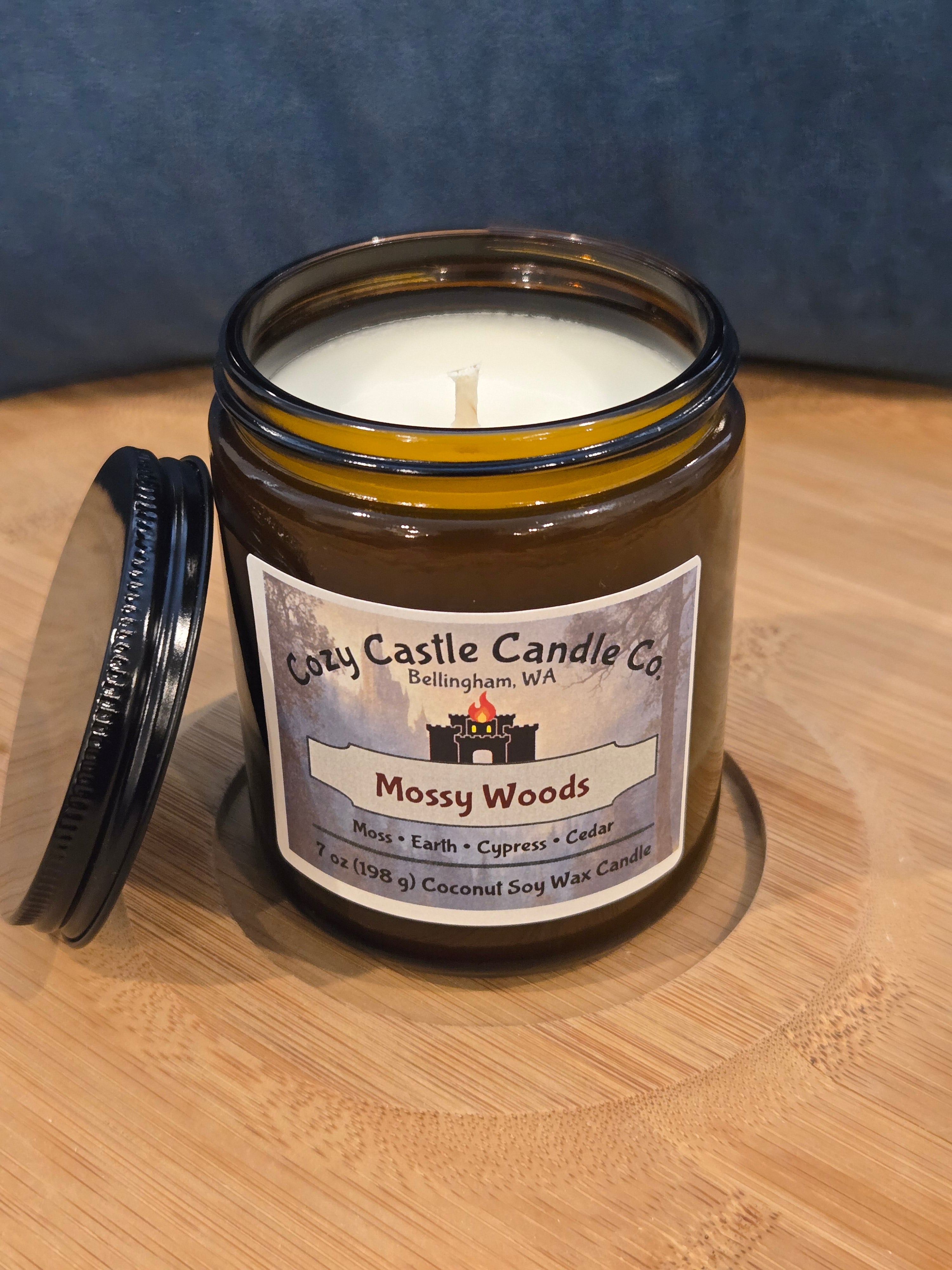 Candle in a glass jar with a label on a wooden surface. Mossy Woods, with notes of Moss, Earth, Cypress and Cedar.