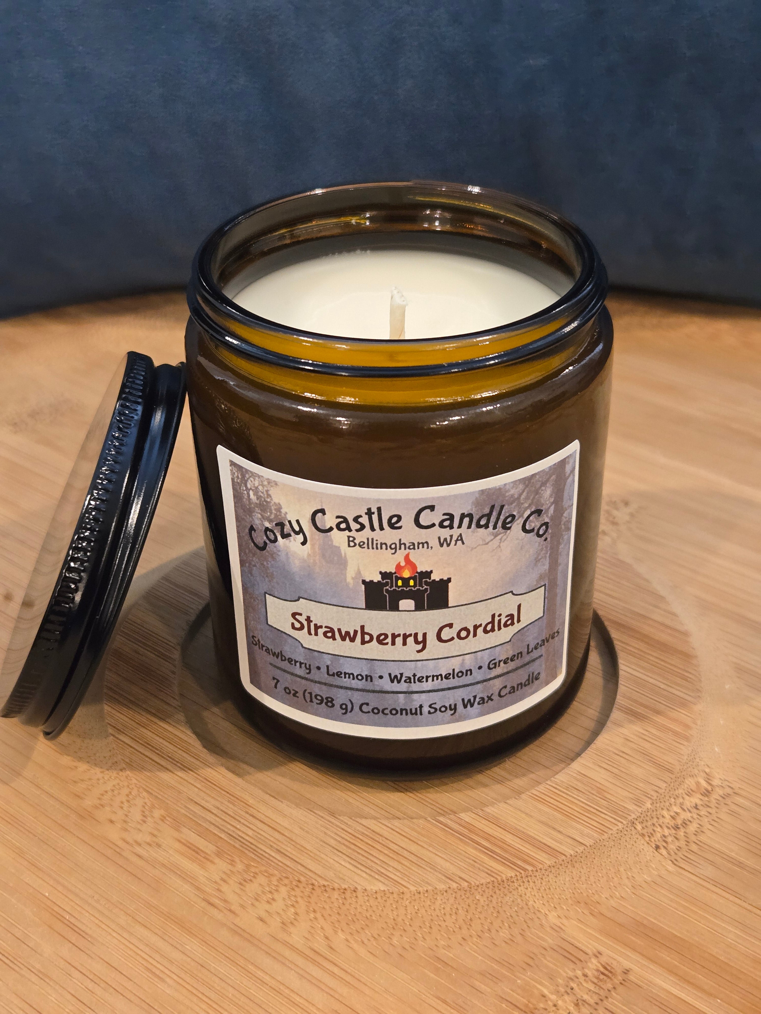 Jar candle with a label on a wooden surface. Strawberry Cordial, with notes of Strawberry, Lemon, Watermelon, and Green Leaves.